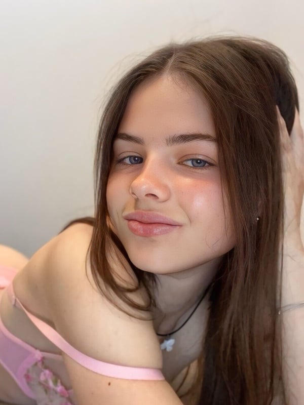 Young woman with long brown hair smiling at the camera while lying down and wearing a pink top and necklace against a plain background.