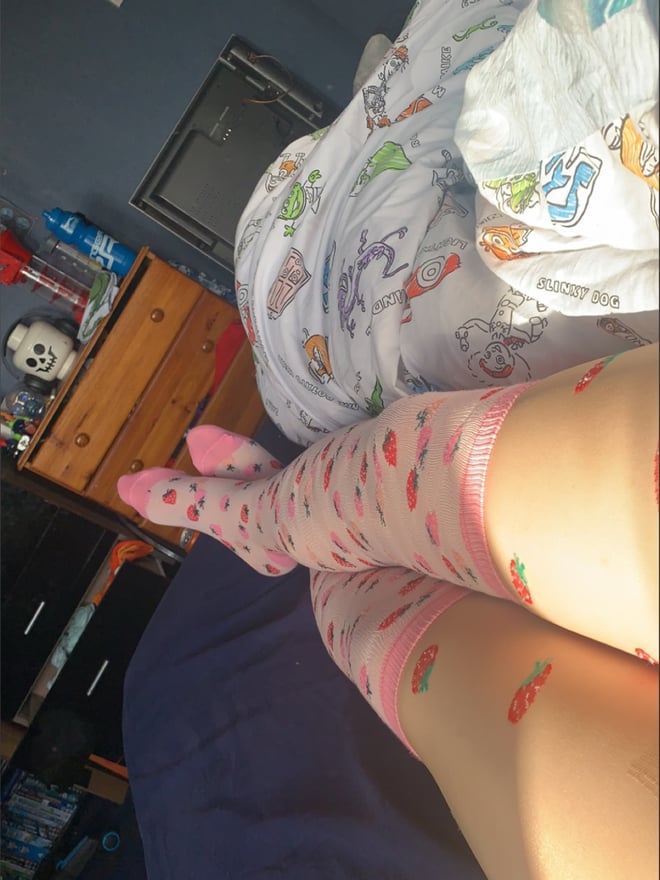 Close-up of legs wearing colorful patterned stockings on a bed in a casual bedroom setting with toys and posters.