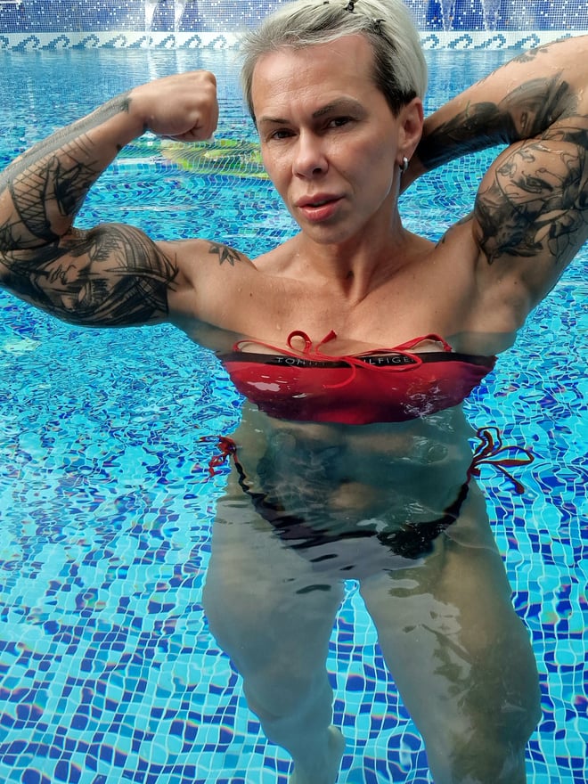 Muscular tattooed woman with blonde hair flexing her biceps in a red bikini top at a swimming pool.