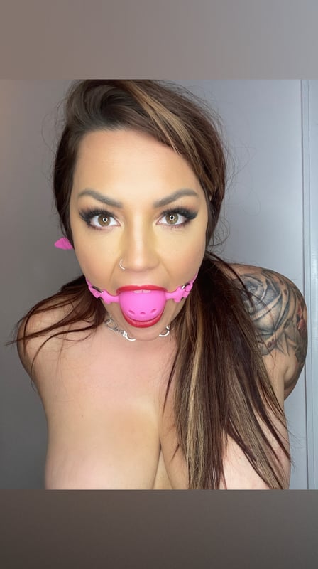 A woman with long hair wearing a pink ball gag accessory around her neck, posing confidently with visible tattoos on her shoulder.