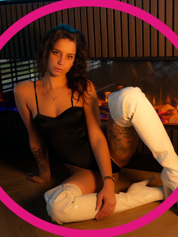 A woman with long dark hair sitting on the floor in a black outfit and white boots in front of a fireplace, enclosed in a pink circle.