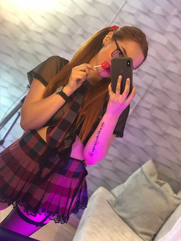 Young woman with glasses and ponytail holding a lollipop and phone, dressed in a sheer top and plaid skirt against a textured wall.