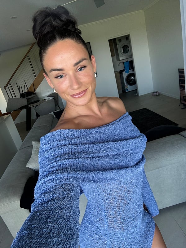 Woman with hair in a bun smiling at the camera, wearing a blue off-shoulder top inside a modern home with casual indoor decor.