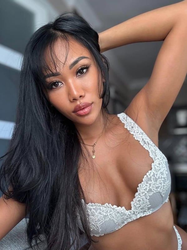 A woman with long dark hair posing confidently indoors, wearing a white lace top, with large windows in the background.