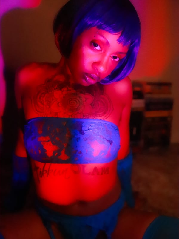 Woman with short blue wig and tattoos posing in colorful lingerie under red lighting.
