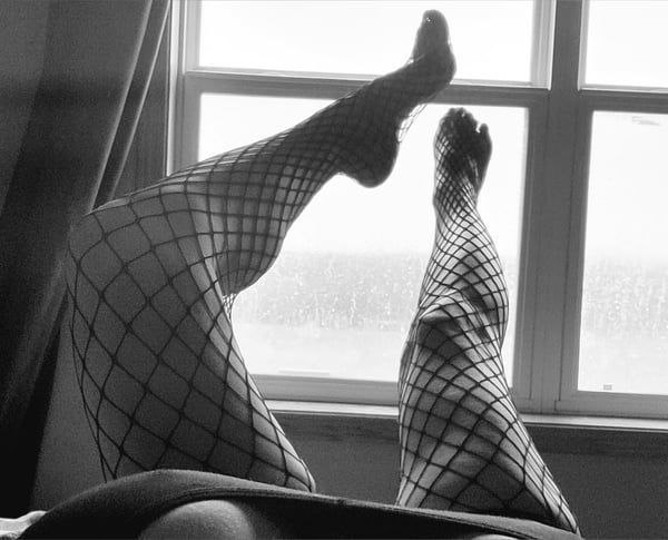 Black and white photo of a woman reclining on a bed with legs raised, wearing patterned stockings, positioned near a window with curtains.