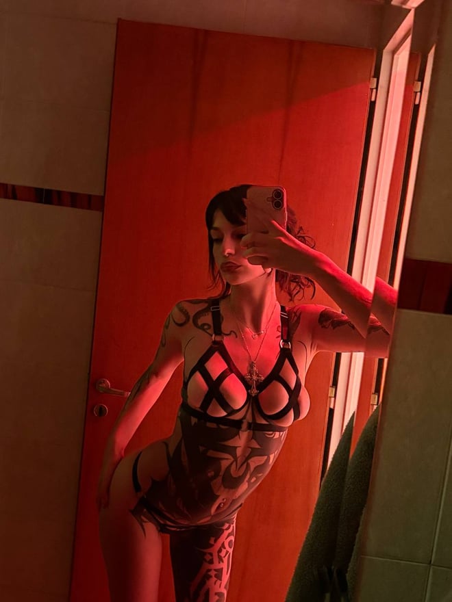 A tattooed woman with dark hair poses in black lingerie and gloves while looking toward the camera.