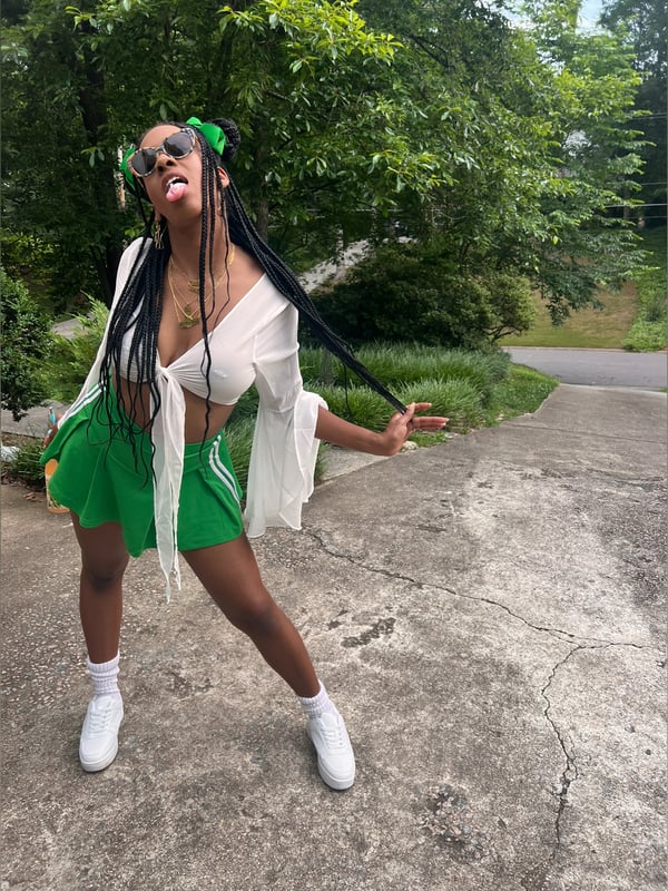 A woman with braids and sunglasses striking a fun pose outdoors in a white top, green skirt, and white sneakers near green trees and a pathway.