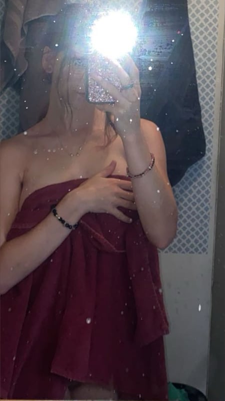 Young woman with blonde hair standing in front of a mirror, wrapped in a red towel, holding her phone for a selfie in a bathroom setting.