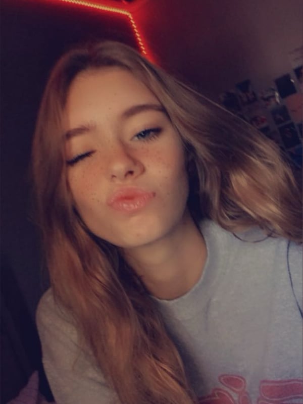 Young woman with long wavy hair making a kissing face, wearing a gray sweatshirt in a bedroom with red LED lights.