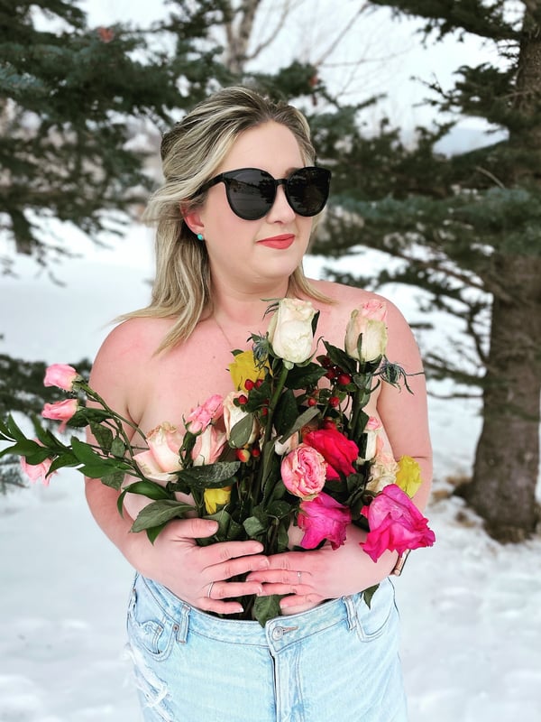 Woman with blonde hair wearing sunglasses and holding a bouquet of colorful roses, standing in snowy woods wearing a light top and jeans.
