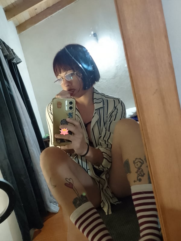 A person taking a mirror selfie in a shirt and striped socks, displaying tattoos on their legs in a cozy indoor setting.