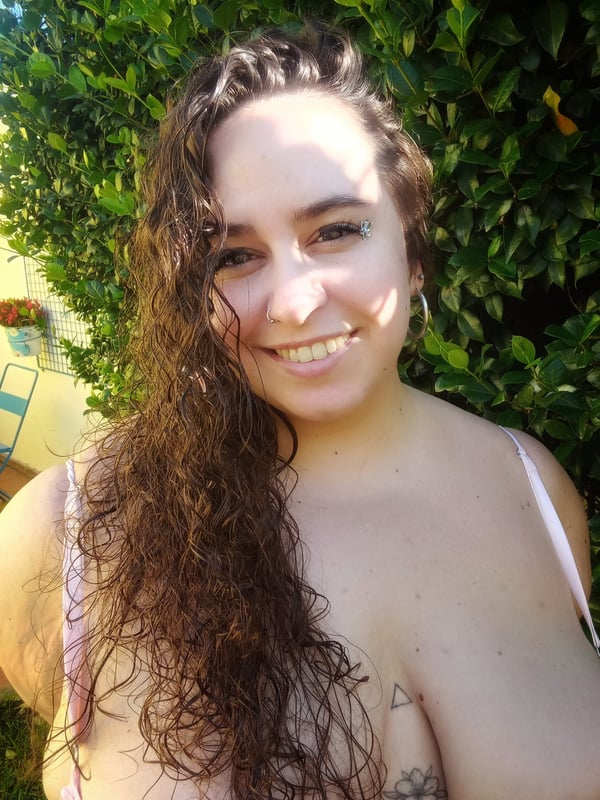 A smiling woman with curly hair posing outdoors in a strap top near green foliage.