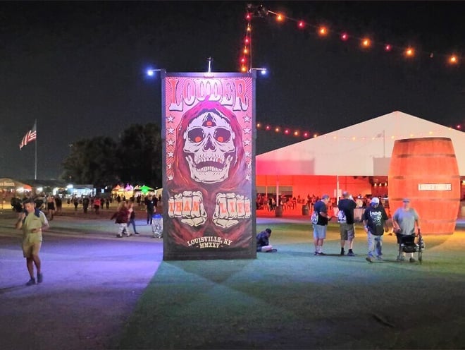 Nighttime festival scene with a large illuminated Løcøs skull banner, colorful lights, an American flag, tents, and people gathered on the grass.