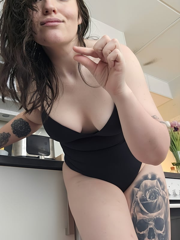 Woman with brunette hair smiling playfully in a kitchen, wearing a black bodysuit and showing tattoos on her arms and legs.