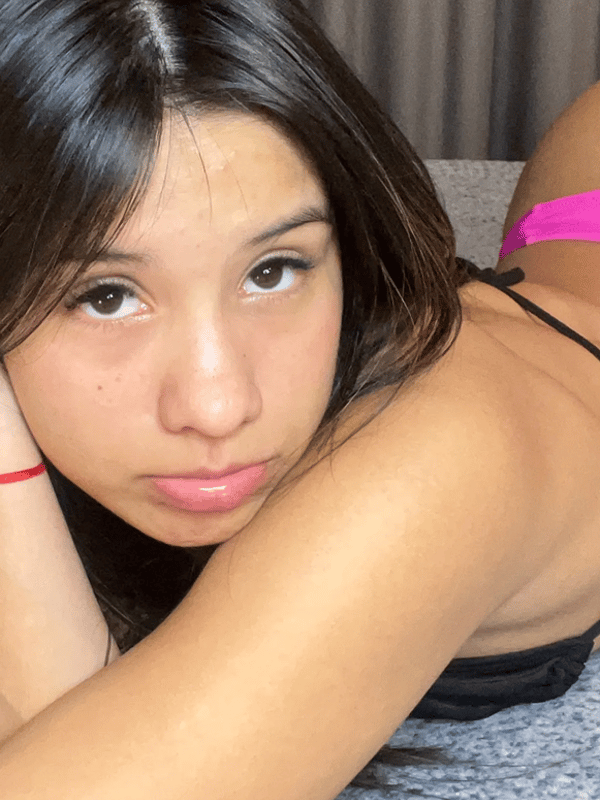 A young woman with long dark hair posing topless while covering her chest with both hands in a bright indoor setting.