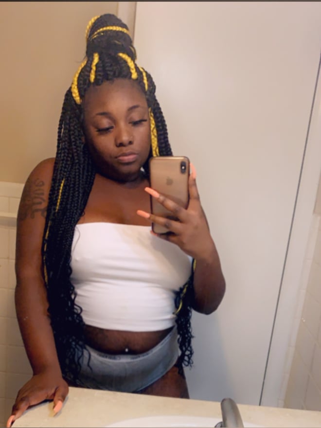 Young woman with long braided yellow hair taking a selfie in a bathroom mirror, wearing a white tube top and gray shorts.