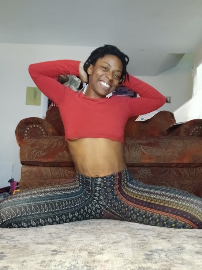 A woman with short curly hair smiling while posing seated on the floor in a red crop top and patterned pants inside a cozy room with a couch.