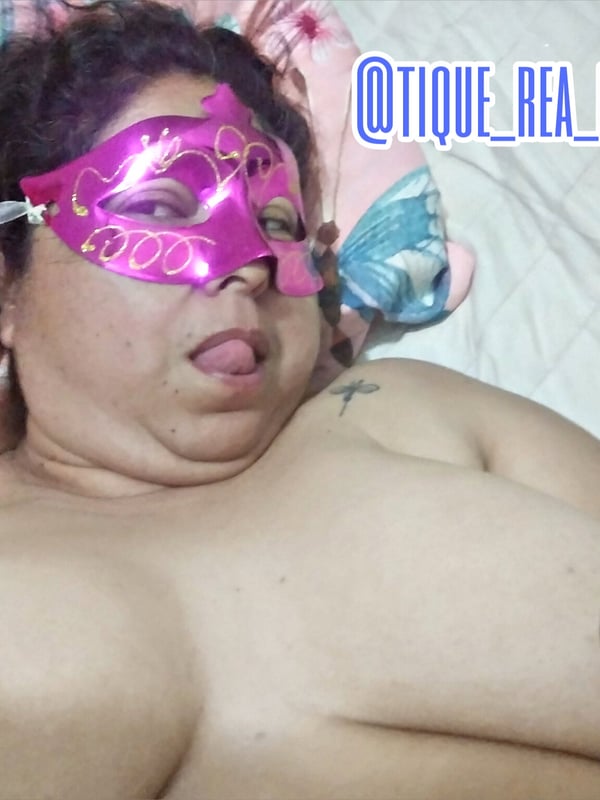 A woman wearing a pink mask and colorful eye accessories, lying on a bed with pillows.