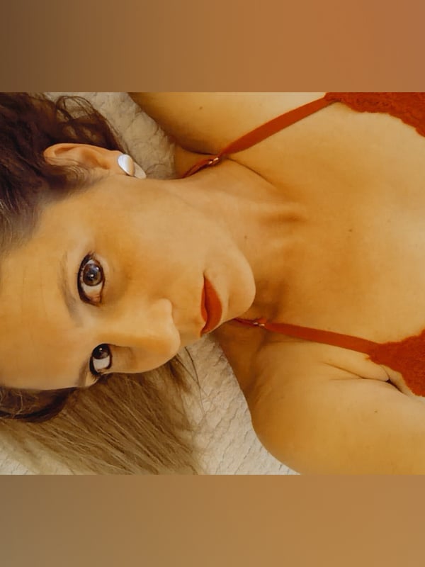 Close-up portrait of a woman with long brown hair wearing a red lace top and jeans, lying down.