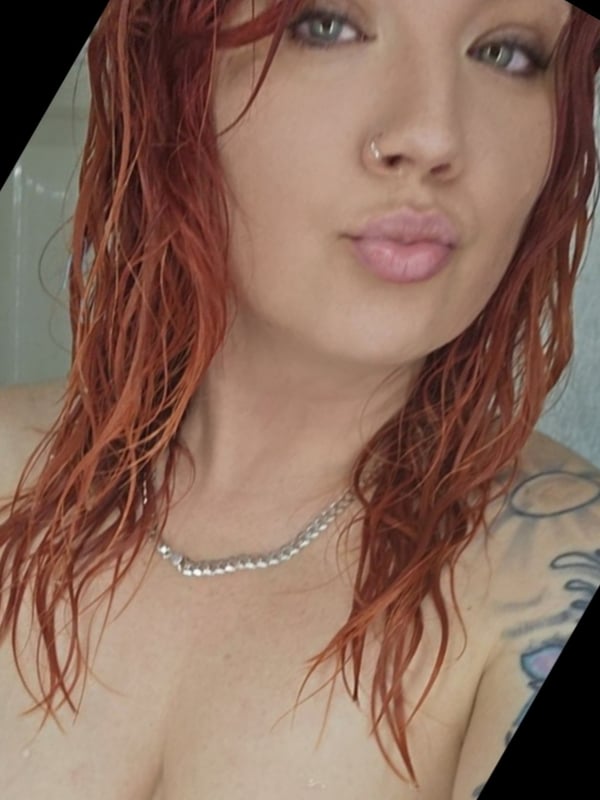 Close-up portrait of a young woman with wet red hair, green eyes, and a nose piercing, wearing a necklace and smiling at the camera.