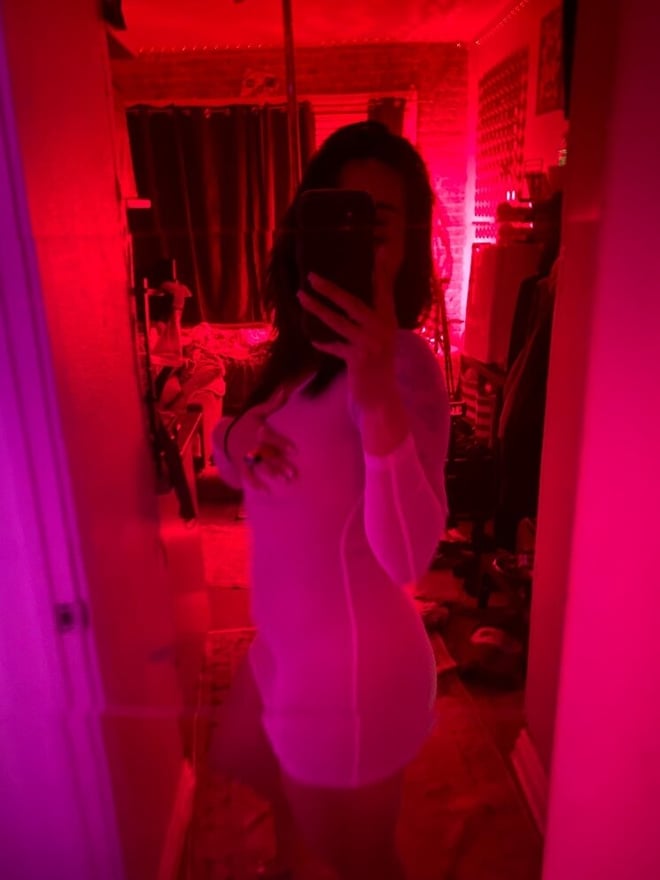 A young woman taking a mirror selfie in a form-fitting white dress in a red-lit room with ambient lighting.