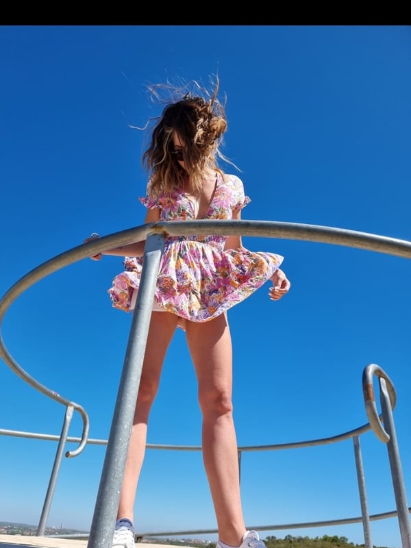 Woman in a floral dress standing on a metal railing against a clear blue sky, with wind blowing her hair and dress.