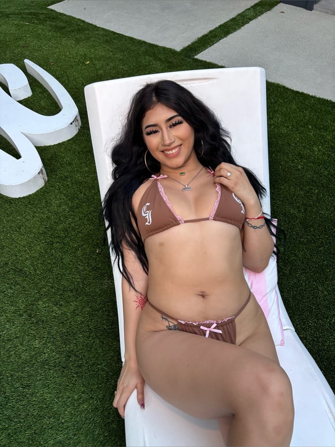 Young woman with long dark hair smiling while lounging on a white lounge chair in a pink bikini on a grassy patio.