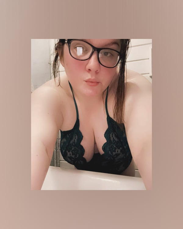 Young woman with glasses wearing a black lace top, posing playfully indoors near a window.