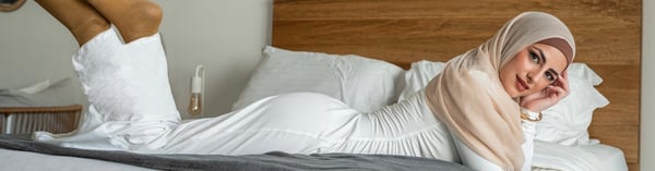 A woman in a hijab and form-fitting white outfit reclines on a bed with pillows and a wooden headboard in a cozy bedroom setting.