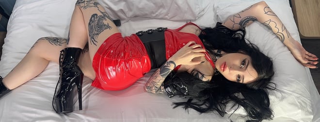A woman with long dark hair and tattoos posing on a bed in a red outfit and black boots.