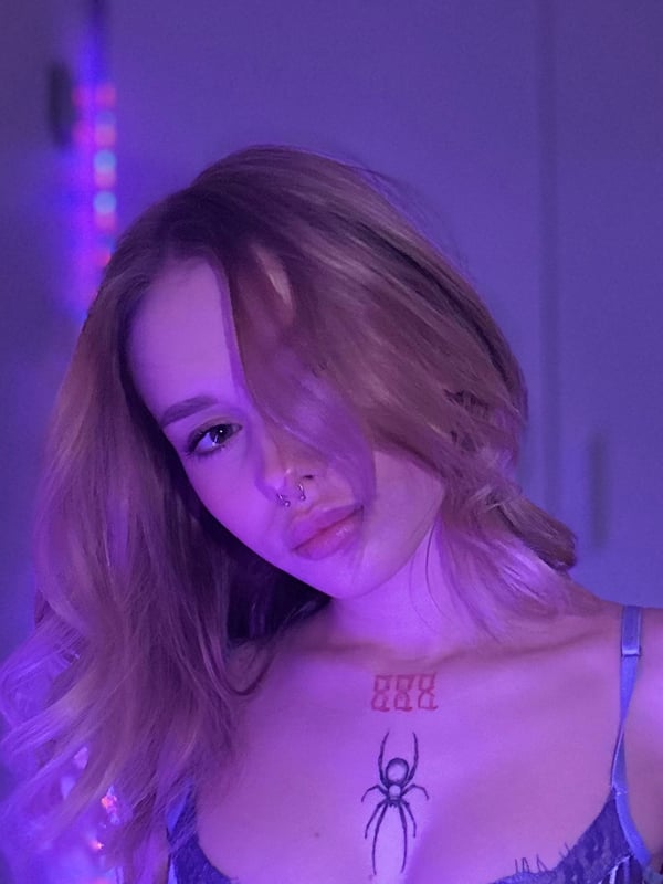 Close-up portrait of a young woman with long blonde hair, a spider tattoo on her chest, and neon lighting in the background.