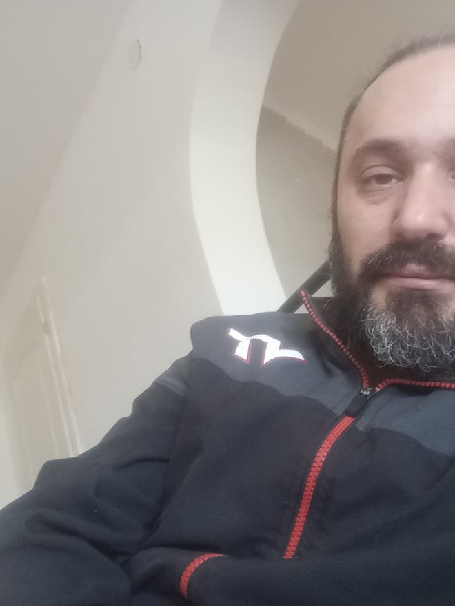 A bearded man with short hair wearing a dark jacket, sitting indoors with a neutral expression.
