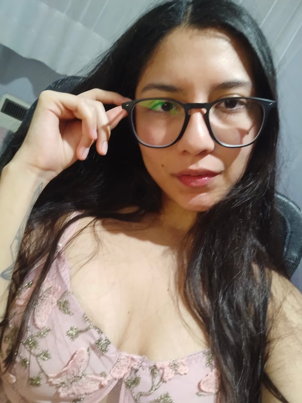 A young woman with glasses and long dark hair smiling at the camera, wearing a floral top while seated.