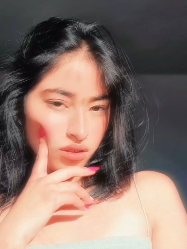 Young woman with long dark hair posing thoughtfully with hand on cheek, wearing a light-colored strap top in a softly lit environment.