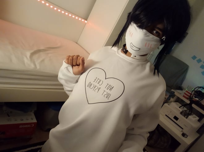 A person wearing a white sweater and face mask, posing in a cozy room with pink ambient lighting.