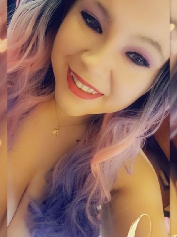 Close-up portrait of a smiling young woman with long pink and purple dyed hair, subtle makeup, and a necklace, against a sparkling background.