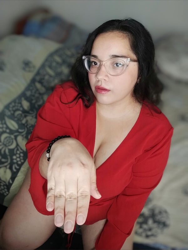 A woman with glasses and long dark hair posing on a bed in a red dress, extending her hand towards the camera.