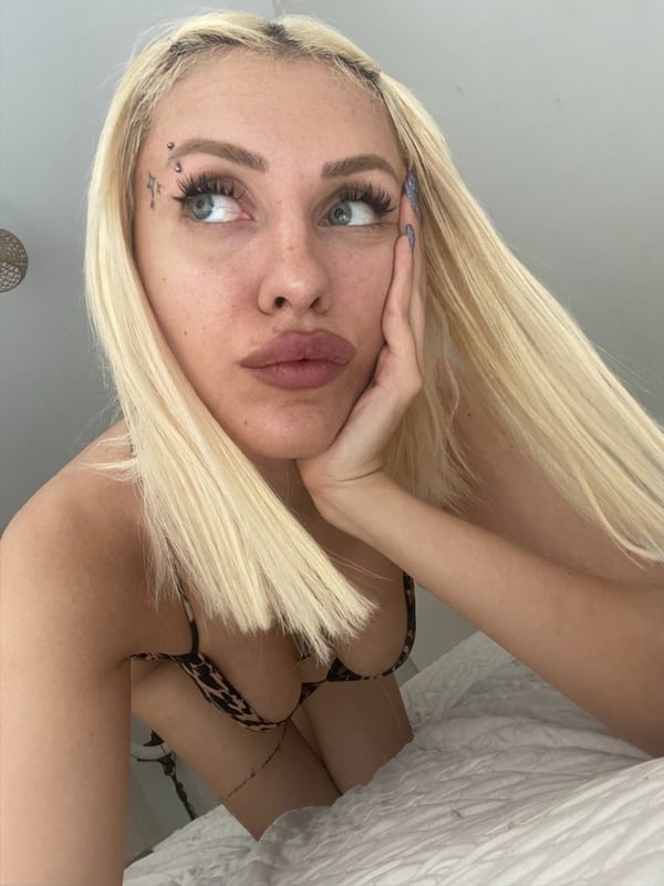 A young woman with long blonde hair and facial piercings posing thoughtfully with her hand on her cheek in a casual indoor setting.
