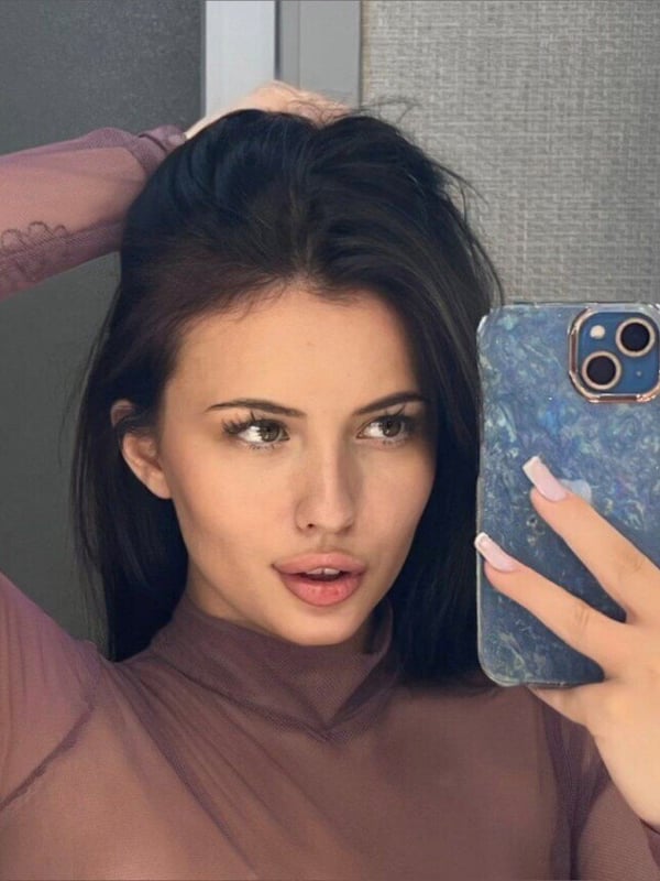 A young woman taking a close-up selfie in a sheer top, holding her phone with a subtle smile.