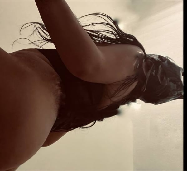 A woman with long dark hair posing sideways in form-fitting black attire in a softly lit room.