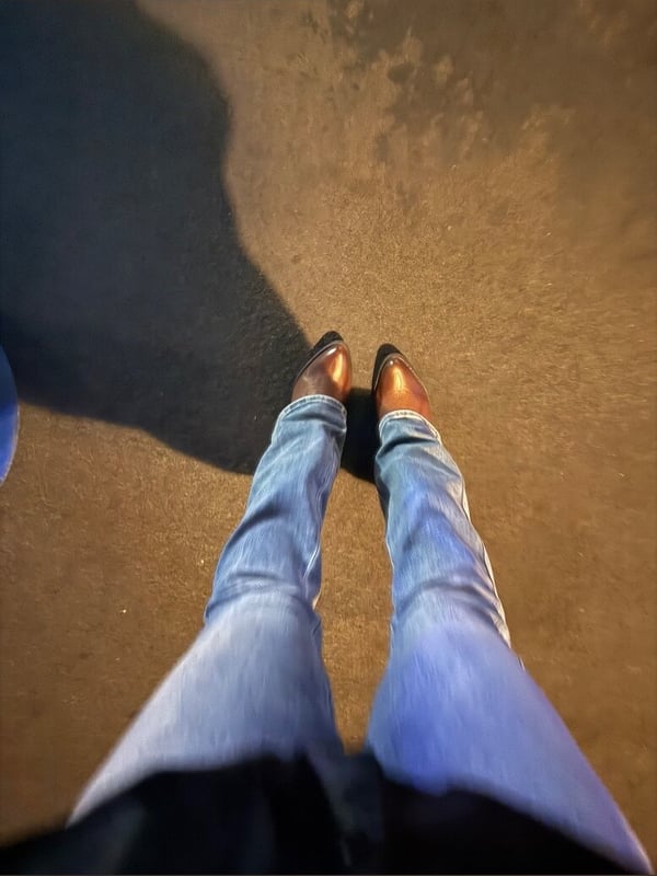 Close-up view from above of a person's legs in blue jeans and brown shoes standing on a sunlit pavement with shadows.