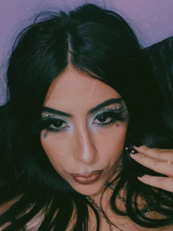 Close-up portrait of a young woman with long dark hair, dramatic eye makeup, and glossy lips posing against a purple background.