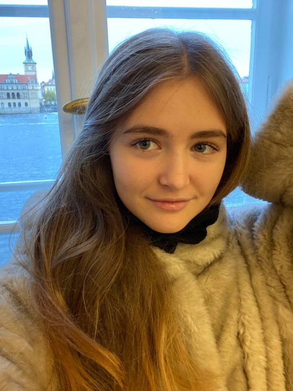 Young woman with long brown hair smiling warmly, wearing a fluffy beige coat, posing by a window overlooking a historic European cityscape with a river and architecture.
