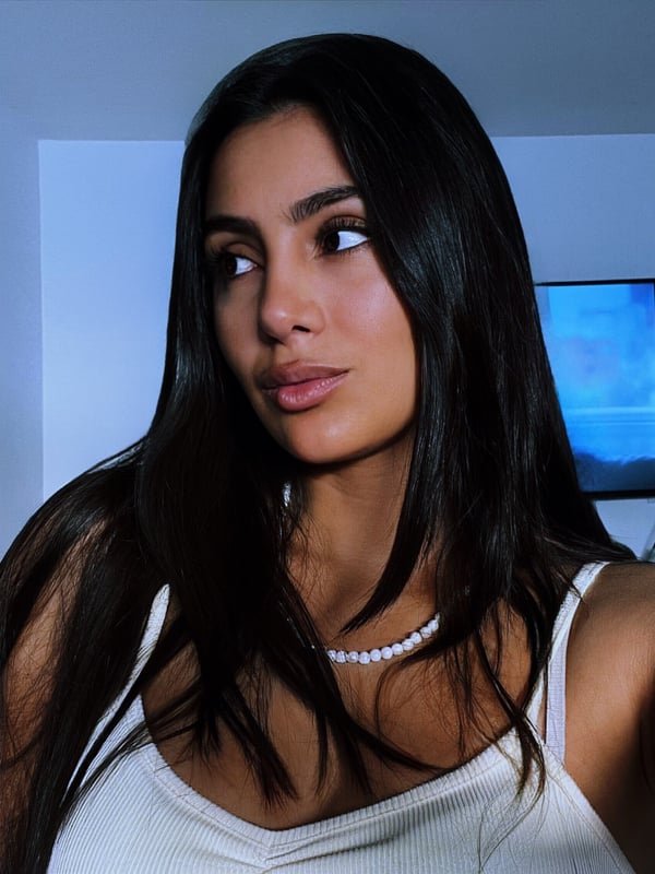 A young woman with long dark hair smiling at the camera, wearing a white top and pearl necklace in a softly lit room.
