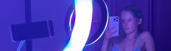 Young woman with blonde hair taking a selfie in front of a ring light, wearing a thin-strapped top in a purple-lit room.