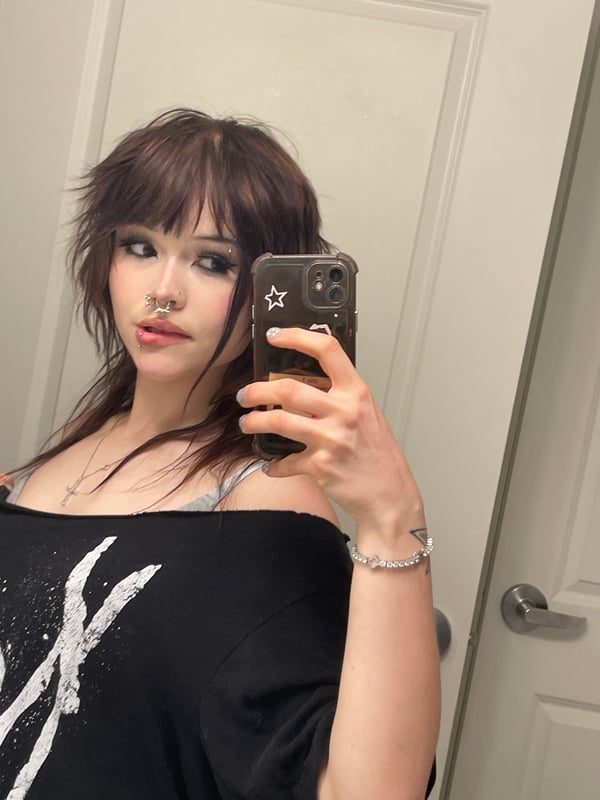 Selfie of a person with brown hair and bold makeup wearing a black lace top and a graphic jacket.
