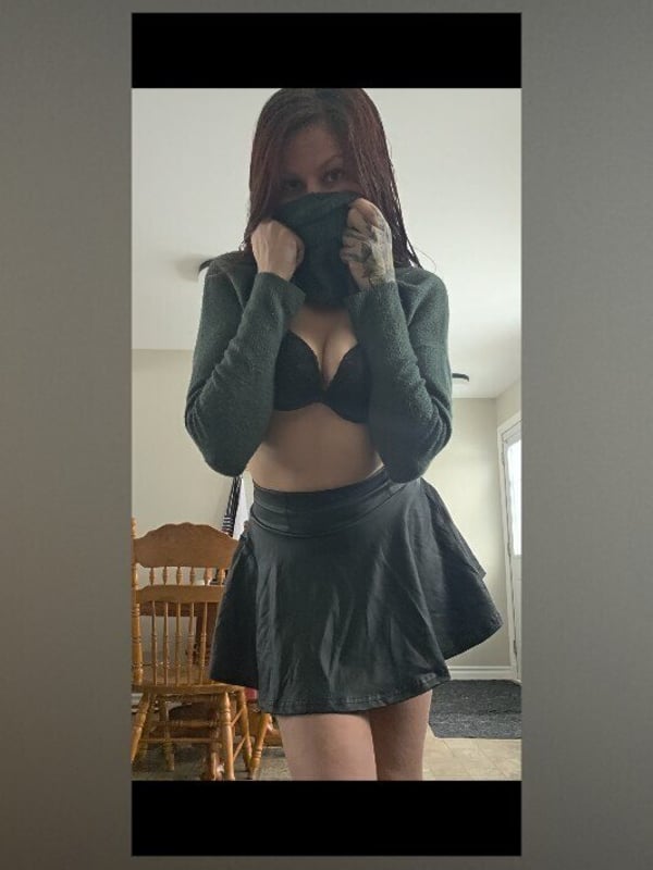 A woman with red hair wearing a green hooded top and black skirt, standing confidently indoors near a wooden chair.