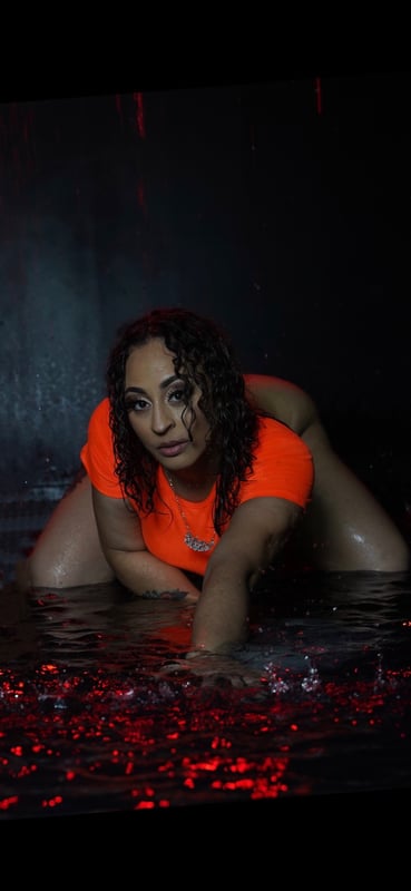 A woman with curly hair posing on the floor in an orange top amidst red petals and water on a dark background.