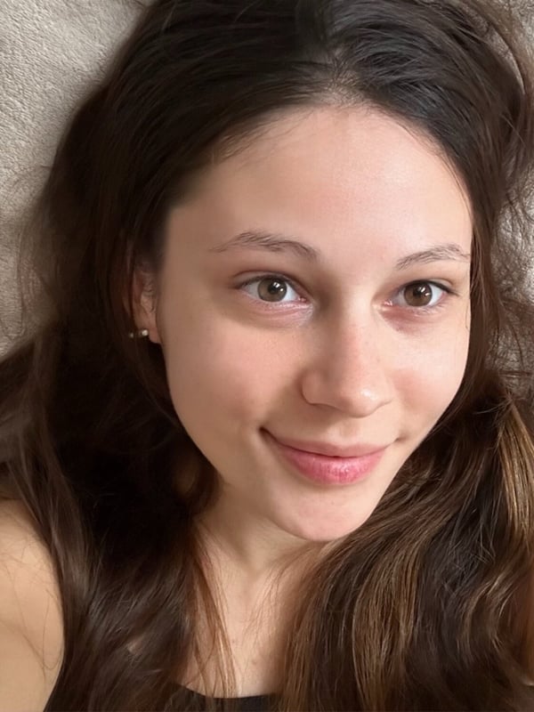 Close-up selfie of a young woman with long brown hair lying on a light-colored surface, smiling softly at the camera.